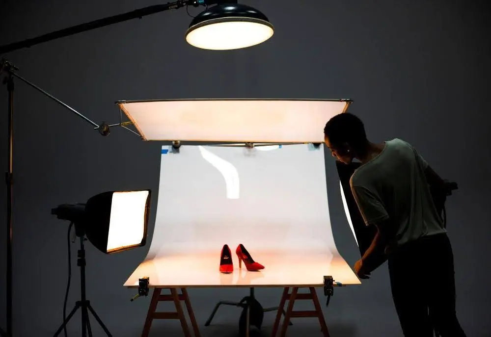 A photographer setting up a product shoot for red high heels, with lighting equipment and a white backdrop in a studio.