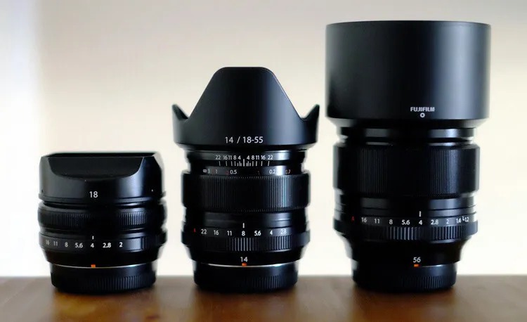 The image shows three Fujifilm camera lenses of varying sizes, placed in a row on a wooden surface.