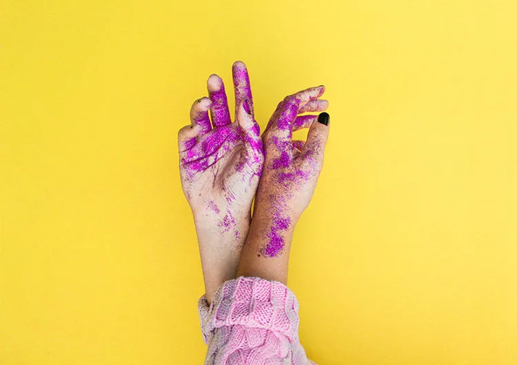 A pair of hands covered in purple glitter, set against a bright yellow background.