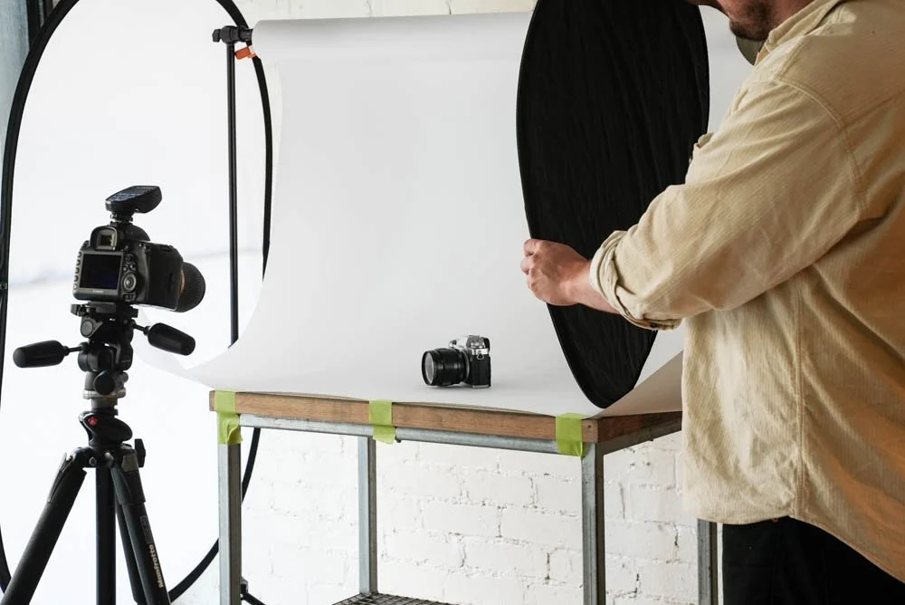 A photographer adjusting a black reflector behind a camera on a white backdrop, and camera adjusted on tripod for product photography.