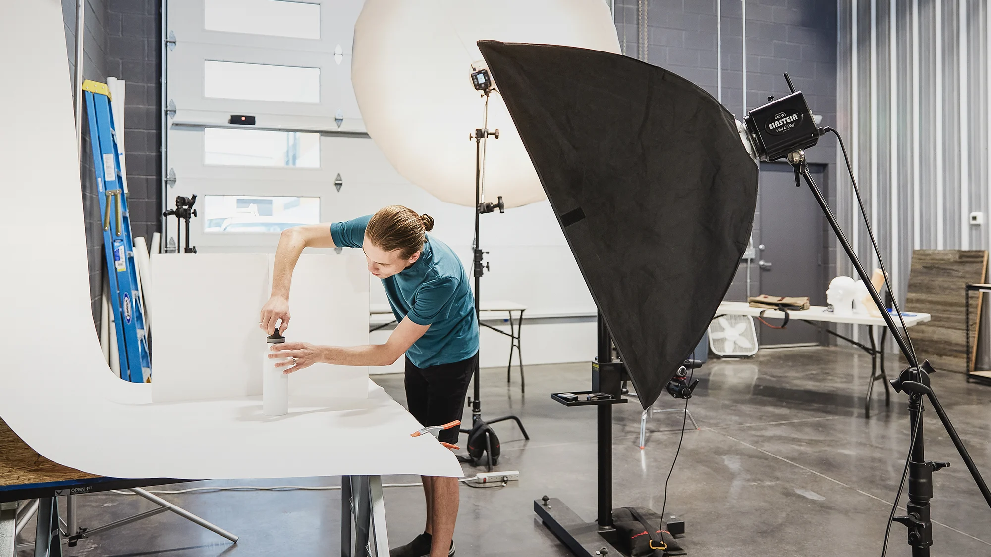 A photographer adjusting a water bottle on a white backdrop with soft lighting and reflectors in a studio setting.