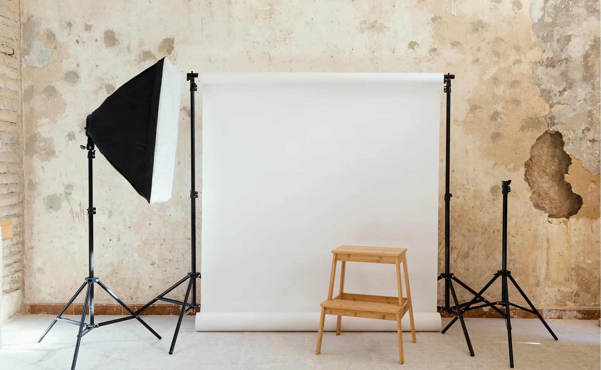 Photography setup with a softbox light, white backdrop, and a wooden stool, set against a weathered wall.