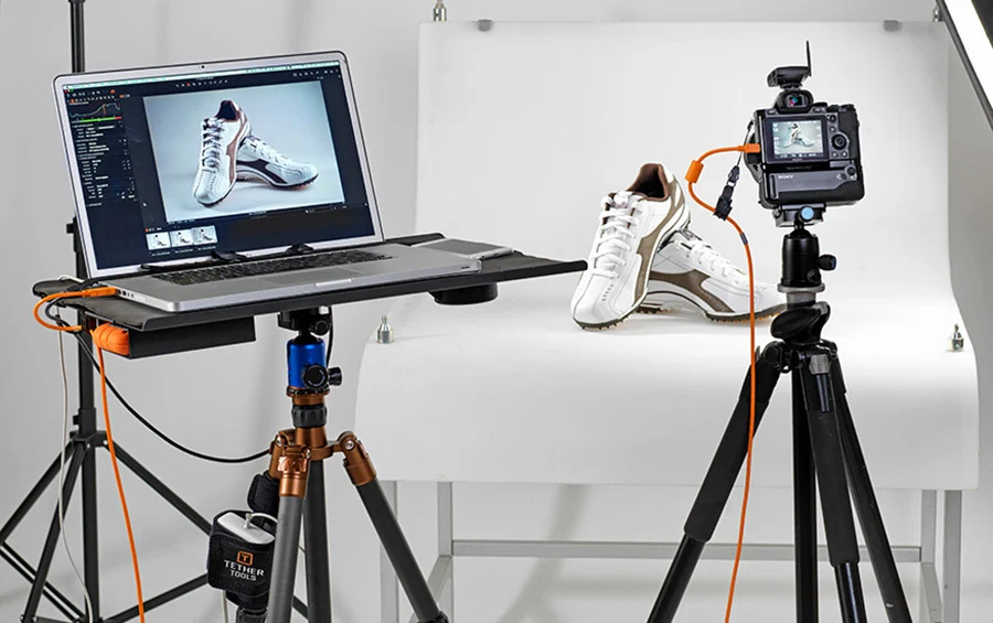 Product Photography setup with a camera on a tripod, a laptop showing the image, and white sneakers on a white backdrop, connected for tethered shooting.