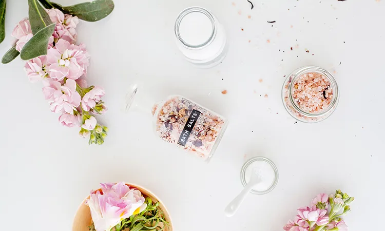 A bird's eye view shot of bath salts, flowers, and jars on a white surface.