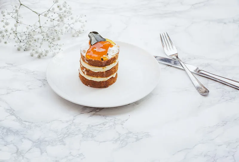 A close-up of a layered cake with cream and a fruit topping, placed on a white plate with a marble background and a sprig of baby’s breath flowers.
