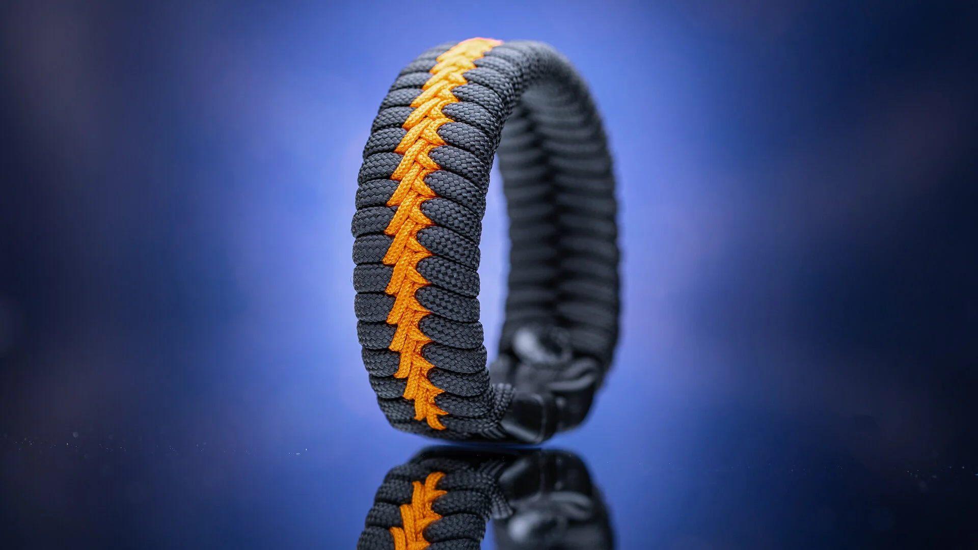 A close-up shot of a black and orange paracord bracelet on a reflective surface with a blurred background.