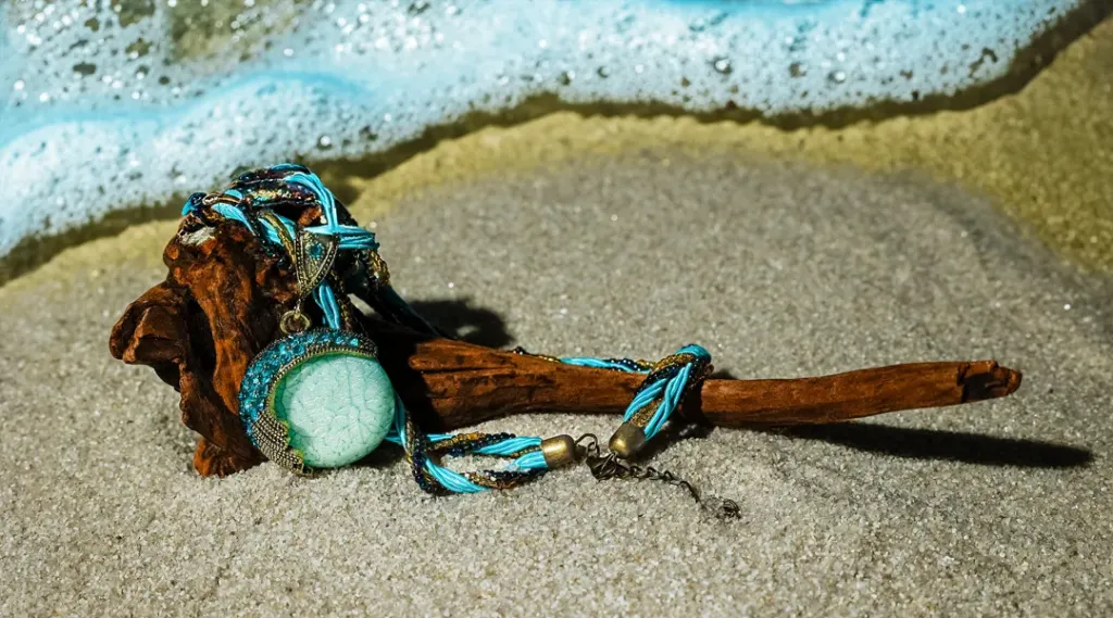 A turquoise beaded necklace draped over a piece of driftwood, resting on sandy beach with ocean foam in the background.