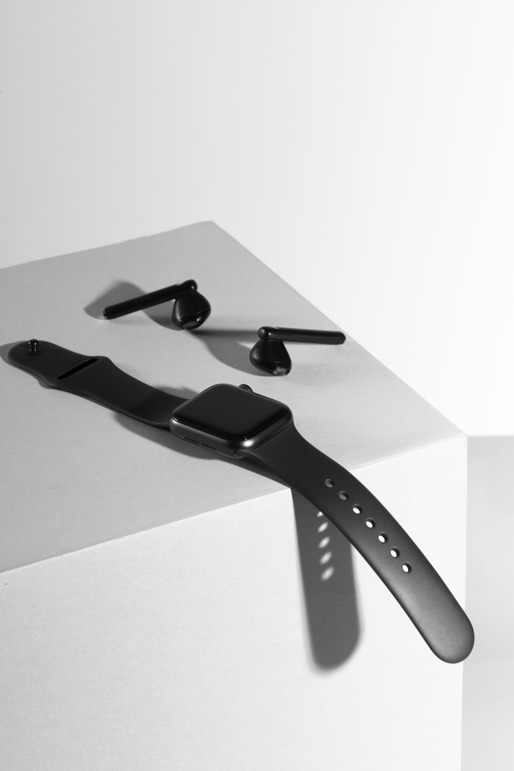 Watch product photography of a black square smartwatch beside wireless earbuds on a white pedestal surface