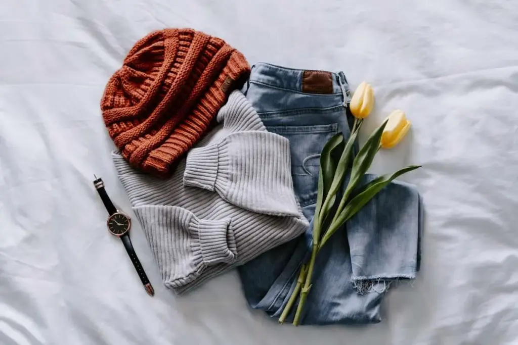 Casual outfit flat lay on a white bed featuring a beanie, sweater, jeans, and tulips