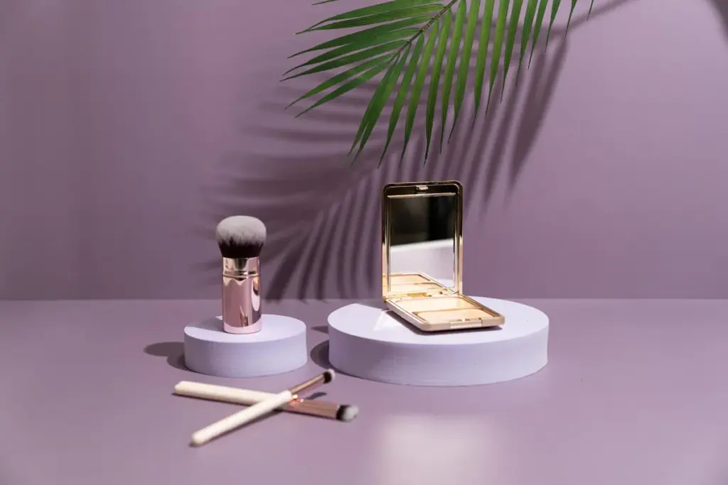 Makeup brushes and a compact palette displayed on white pedestals against a purple backdrop with palm leaf shadows.