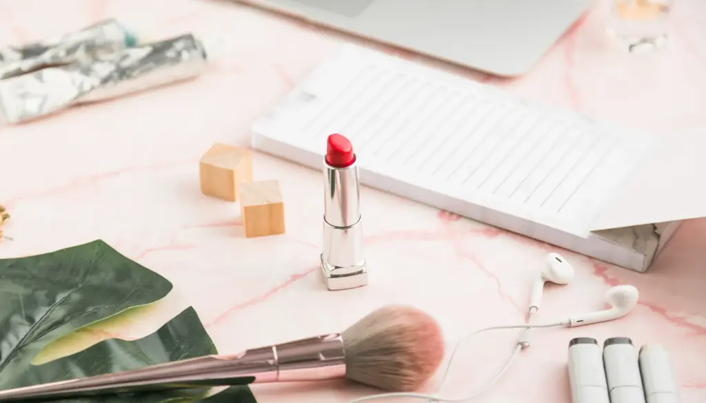 Red lipstick and makeup brush on a pink marble desk with a notebook, earbuds, and decorative leaves.