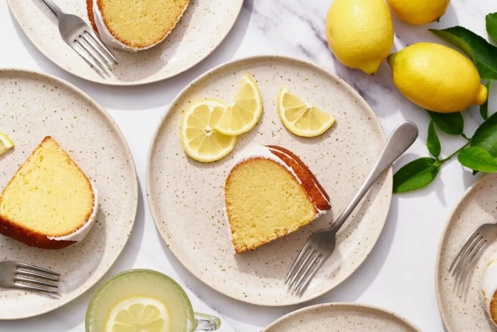 Slices of lemon cake on speckled plates with lemon wedges and a glass of lemonade, styled for food photography.
