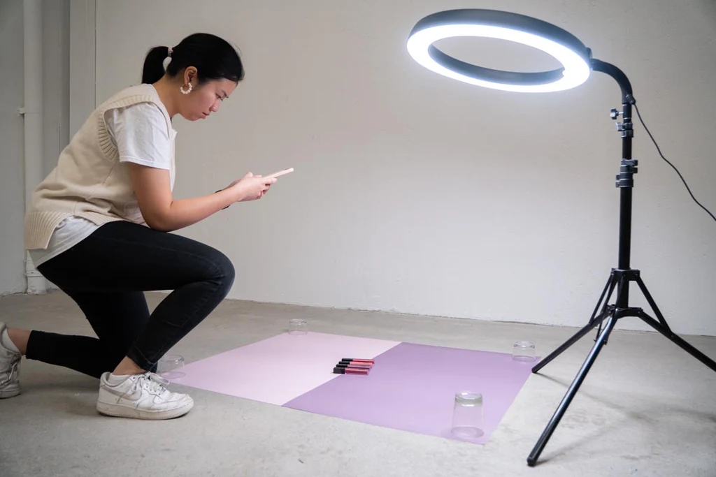 Woman setting up a product photography shoot with a ring light, colored backdrop, and small makeup items.
