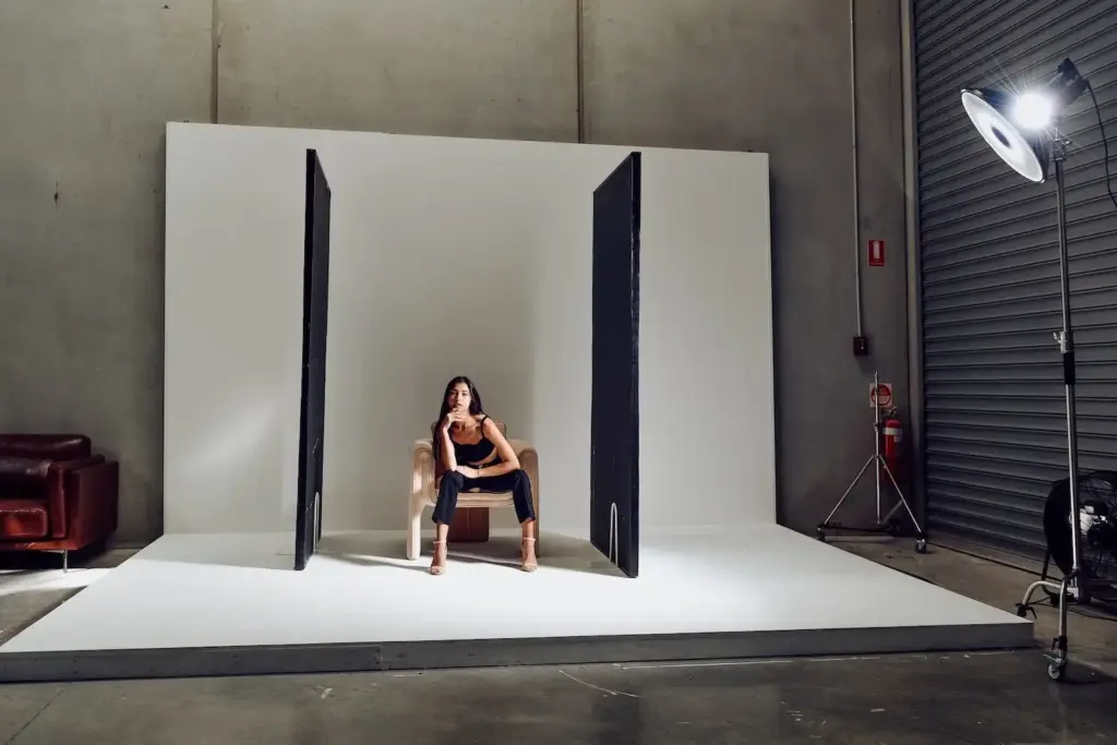 Model seated on a studio set between black polyboards and lighting equipment.